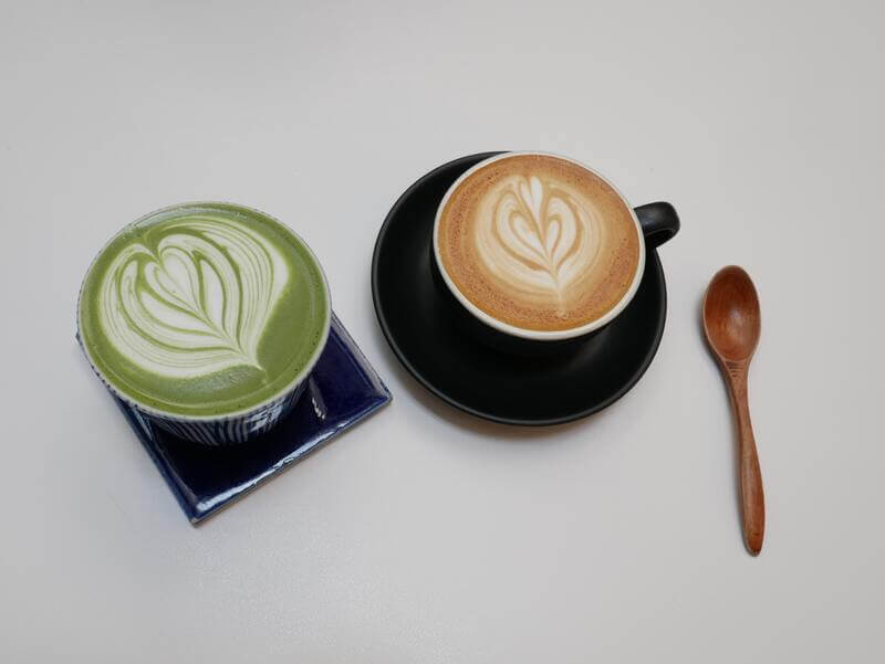Na stole leżą dwie filiżanki – jedna z cappuccino, druga z matcha latte – zdobione sercem na piance.