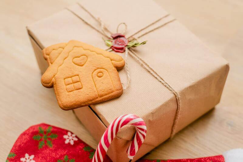 Na stole leży ekologicznie zapakowany prezent w świątecznej otoczce - skarpecie mikołaja, lizaku oraz z domkiem z piernika jako dodatkiem.